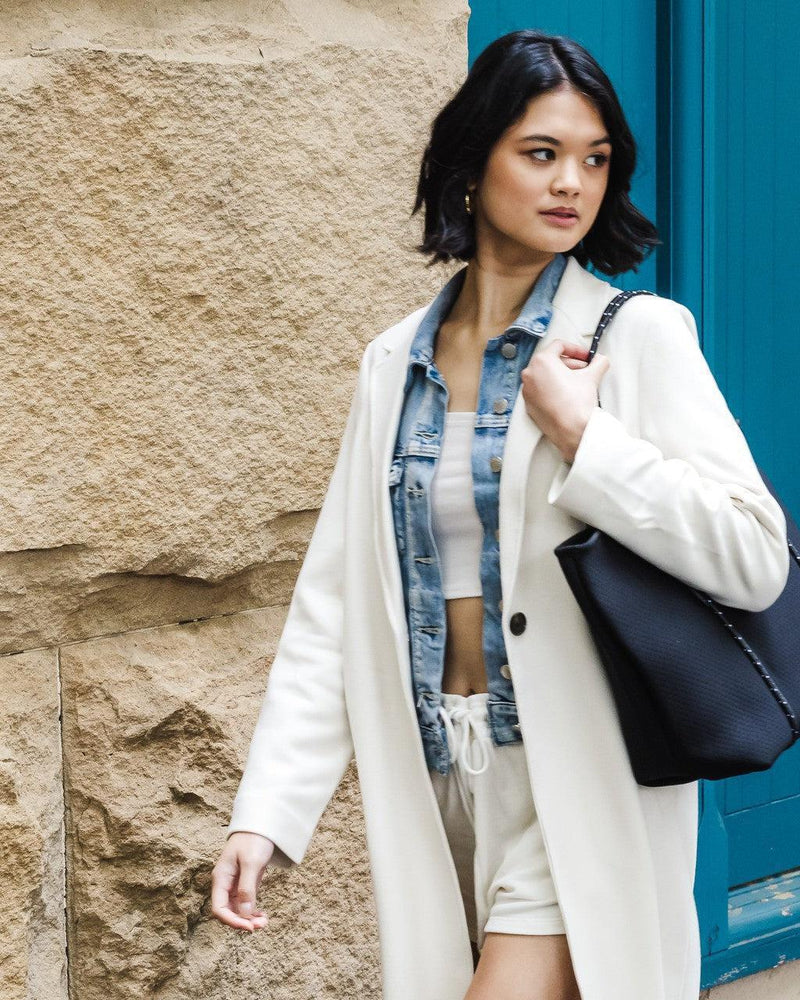 Woman carrying a black perforated neoprene tote bag with studded rope handles, walking past a stone wall.