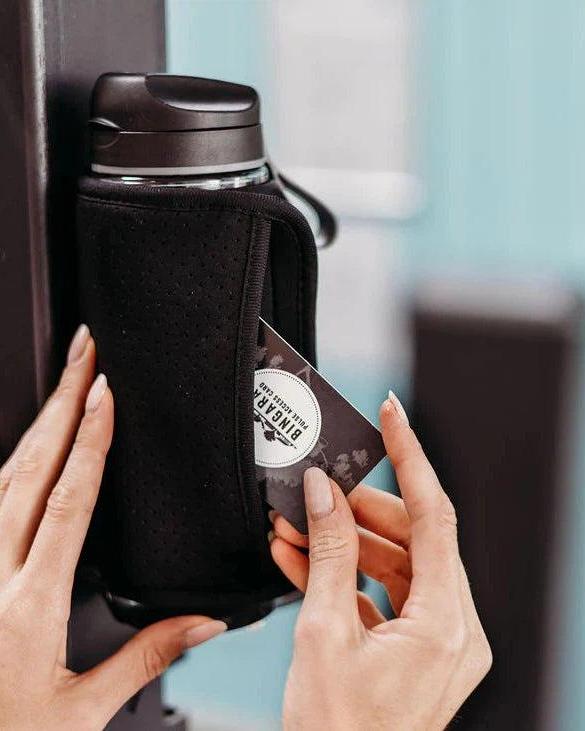 Jet black magnetic gym caddy holding a water bottle, with a hand inserting an access card into its side pocket.