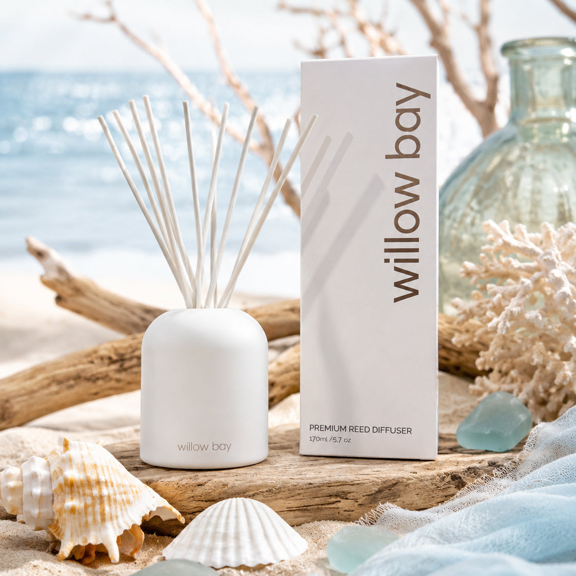 White Willow Bay Premium Reed Diffuser with reeds, packaging, seashells, and driftwood on a beach.