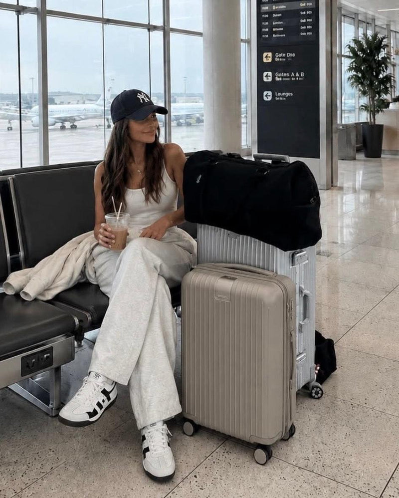 Woman in a white tank top and grey pants sits in an airport with a black Willow Bay Cabin Bag on a silver suitcase.